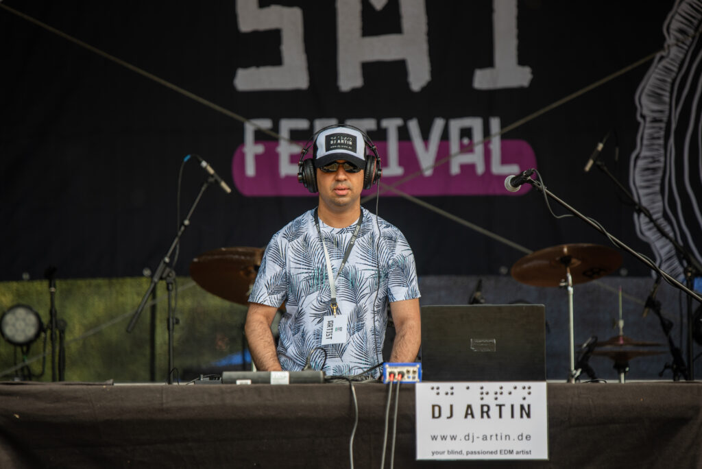 DJ Artin steht auf der Bühne eines Festivals mittig hinter dem DJ Pult.
